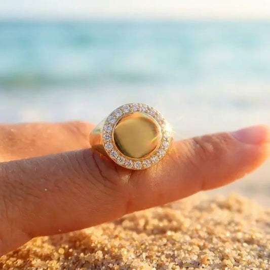 Gold circular signet ring with diamonds on a finger against a beach background