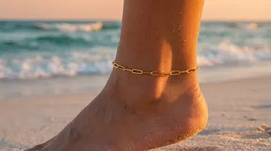 Close up of a foot with a gold paperclip chain on a sandy beach with ocean waves in the background