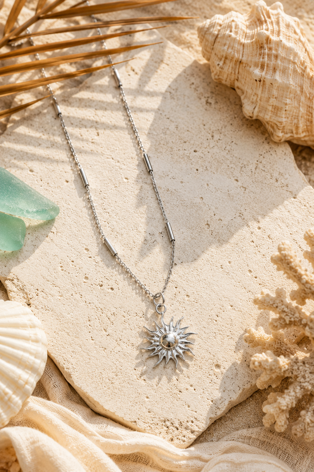 Silver bar station chain with sun pendant lying on a cream-colored surface with seashells and sea glass surrounding it.