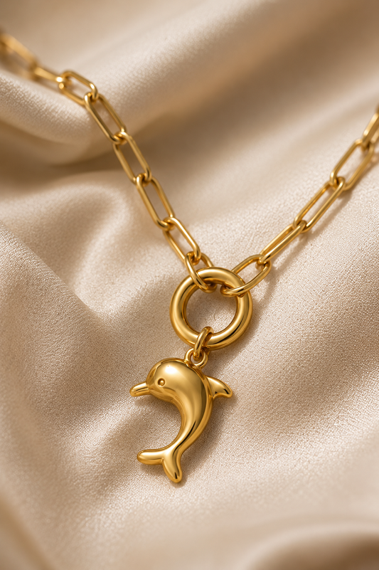 Gold paperclip chain with dolphin pendant lying on cream colored satin.