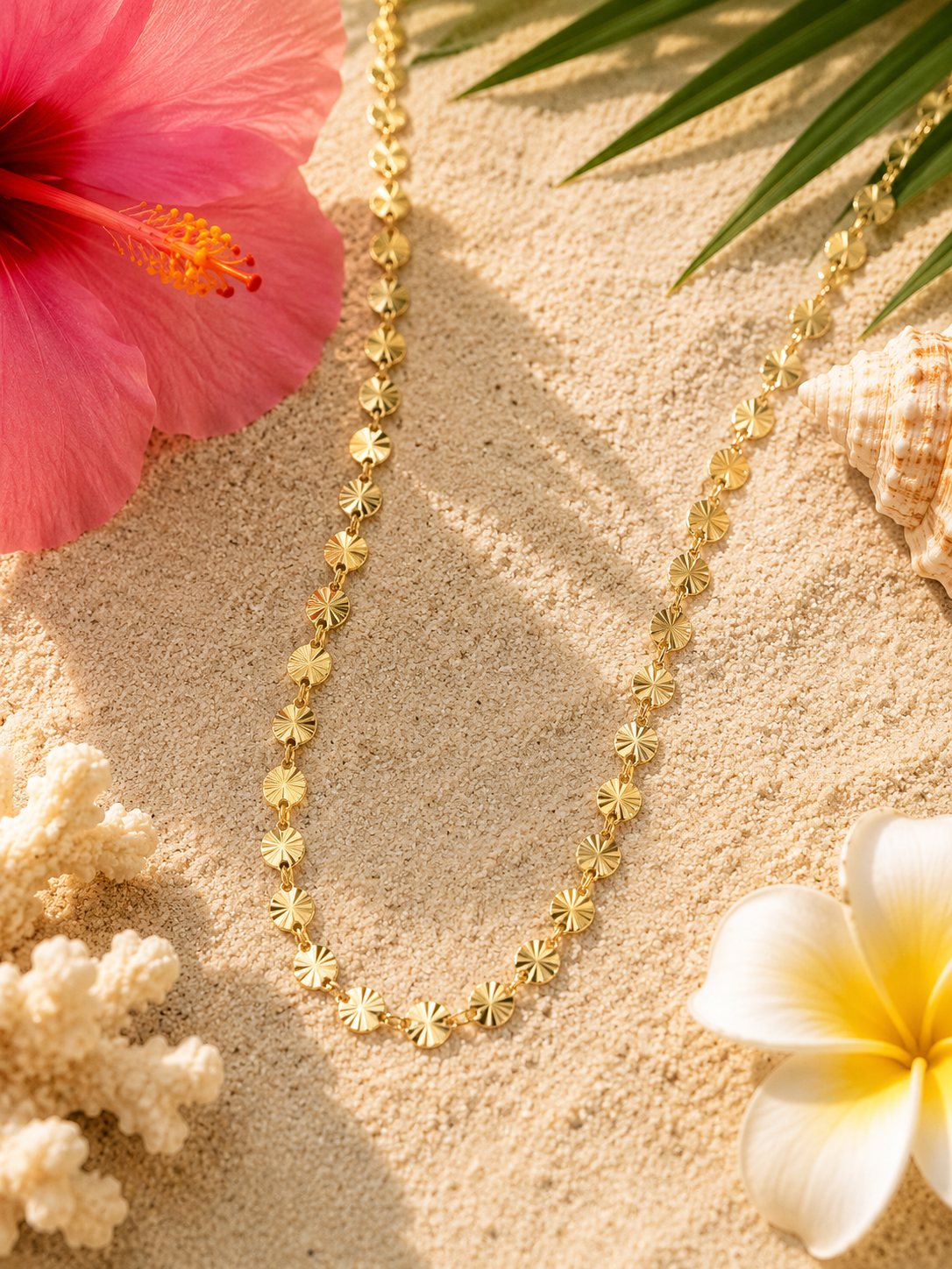 A gold-toned necklace with small sunburst-shaped beads, laid on sandy ground with tropical props including a pink hibiscus flower, a white plumeria flower, coral, and a seashell.