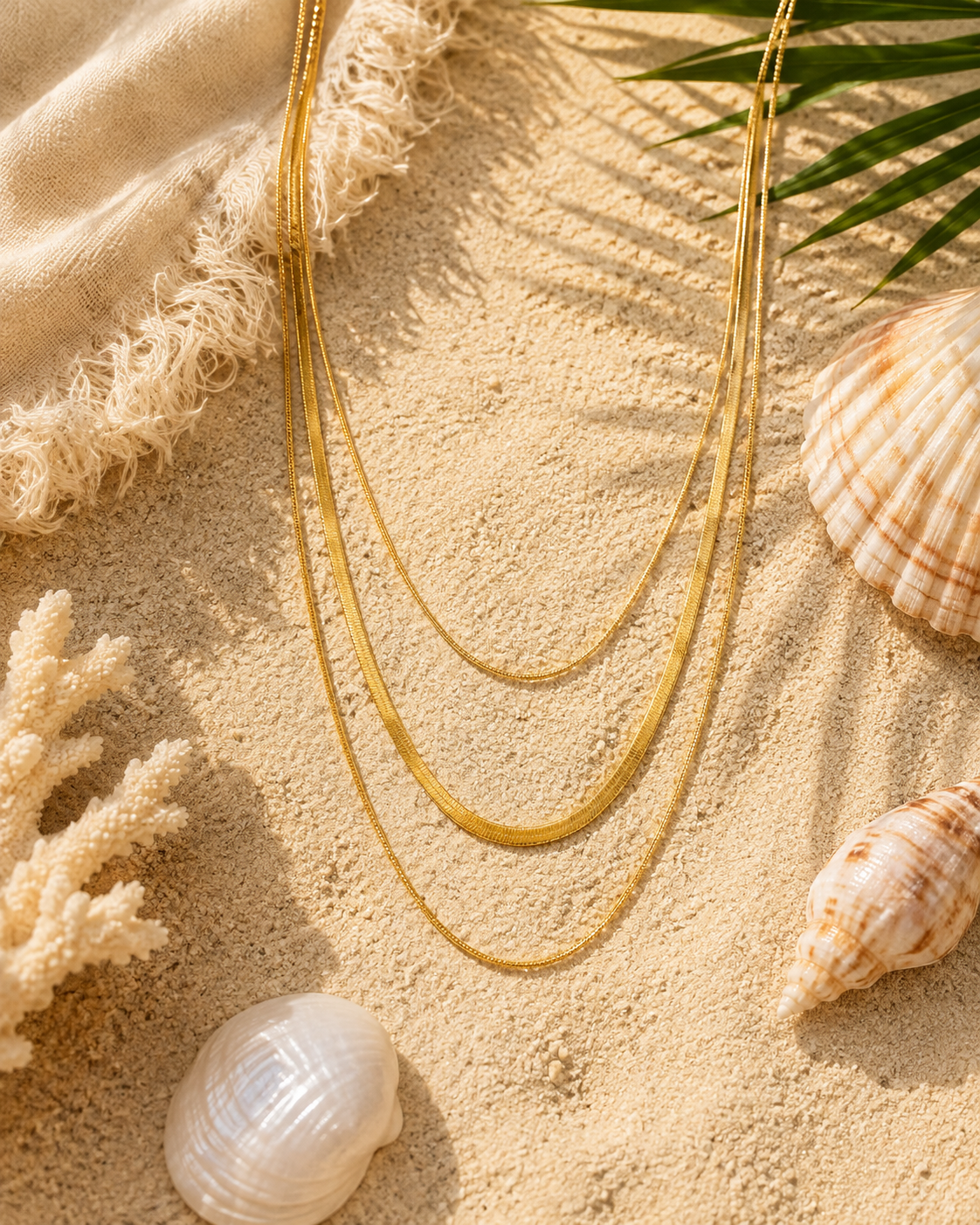 Three layered gold chain necklaces arranged on a sandy beach-style surface with seashells, coral, a fringed textile, and palm leaves.