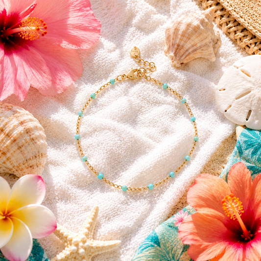 A delicate gold chain bracelet with small turquoise beads, styled on a white towel with seashells, starfish, hibiscus flowers, and a woven straw hat.