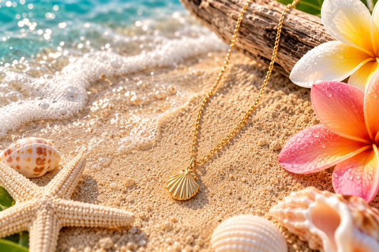 A gold chain necklace with a scalloped shell pendant is displayed on a sandy beach scene with tropical flowers, seashells, a starfish, and ocean water in the background.