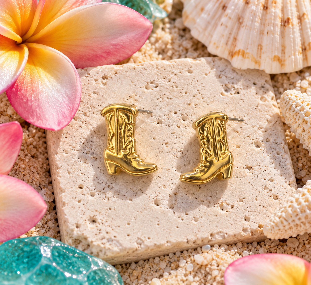 Cowboy boot earrings on a white stone with a plumeria flower and sea shell nearby.
