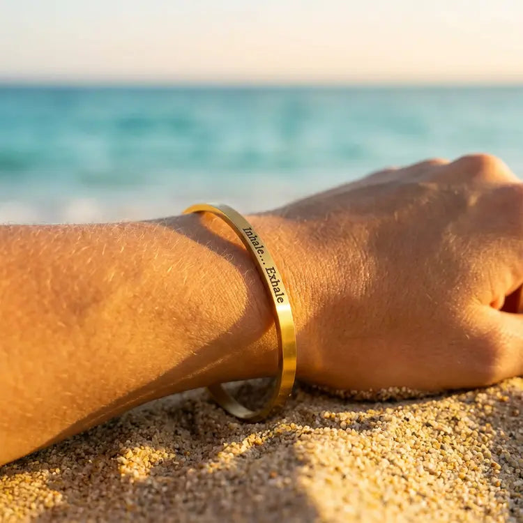 Gold cuff bracelet on a wrist with "Inhale...Exhale" engraved on it with the ocean in the background