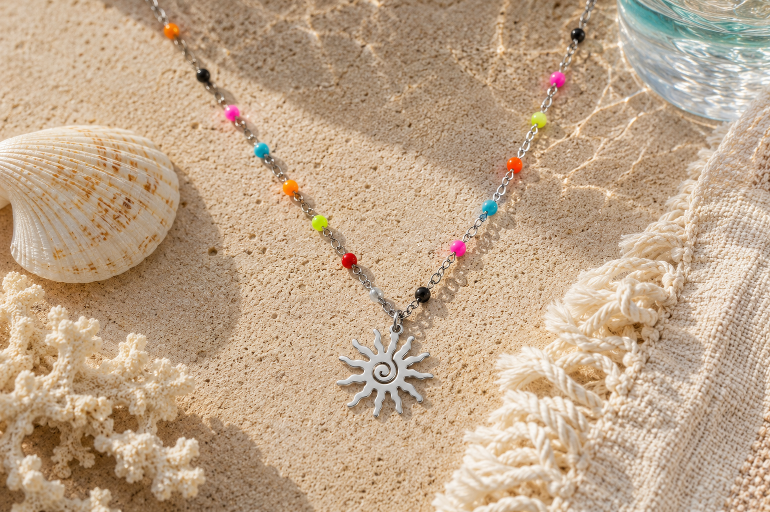 Silver and colorful beaded necklace with spiral sun pendant lying next to a beach blanket, coral and a seashell.