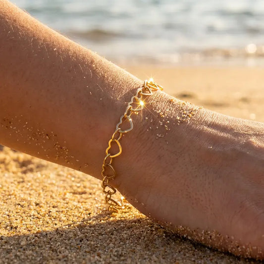 Close up of a dainty gold chain bracelet made of tiny hearts on a person's wrist with a beach background