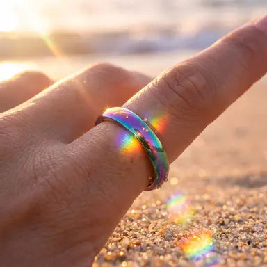 Hand wearing a colorful ring with a rainbow effect on a beach at sunset.