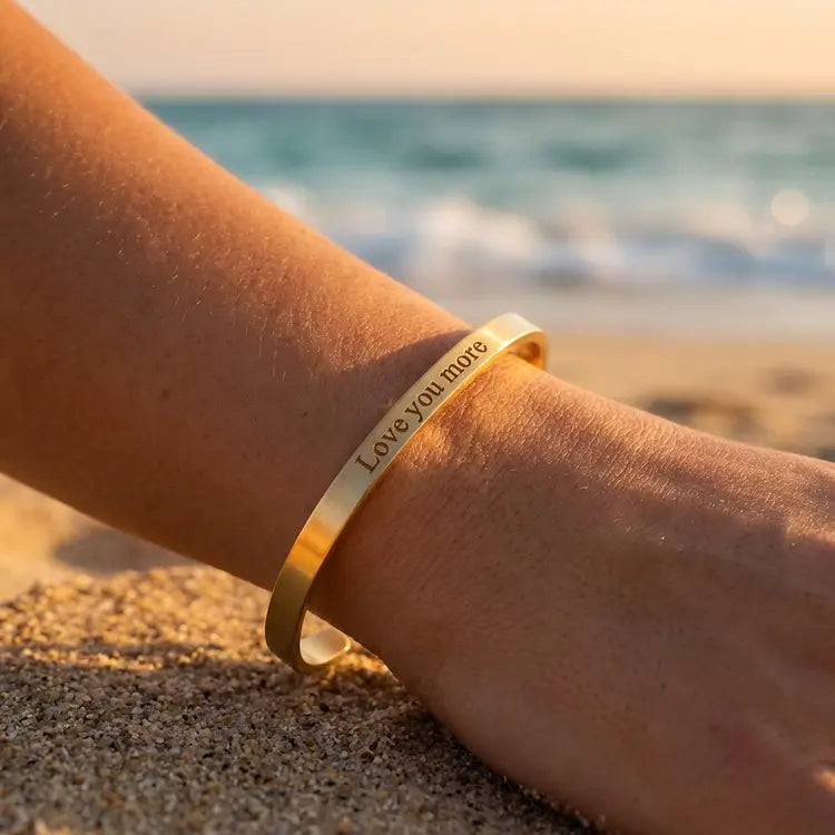 Gold bracelet with 'I love you more' engraving on a person's wrist by the beach.