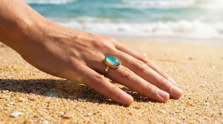 Hand wearing a mood ring with a blue green stone on a sandy beach