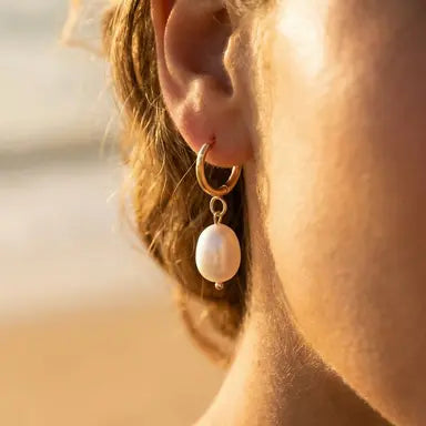 Close-up of an ear wearing a gold hoop earring with a pearl pendant against a blurred background.