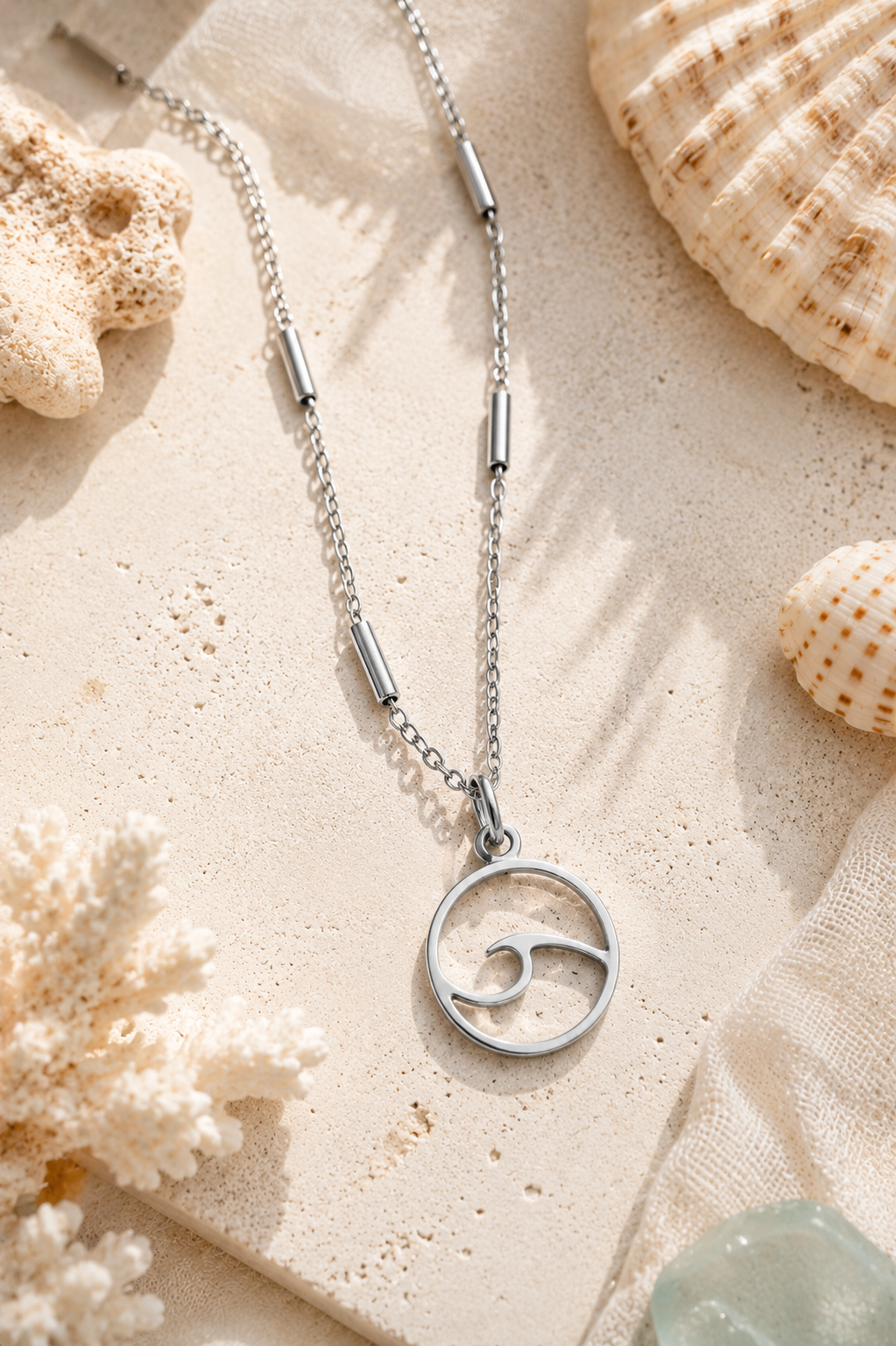 Silver necklace with a circular wave pendant on a sandy background with shells and coral.