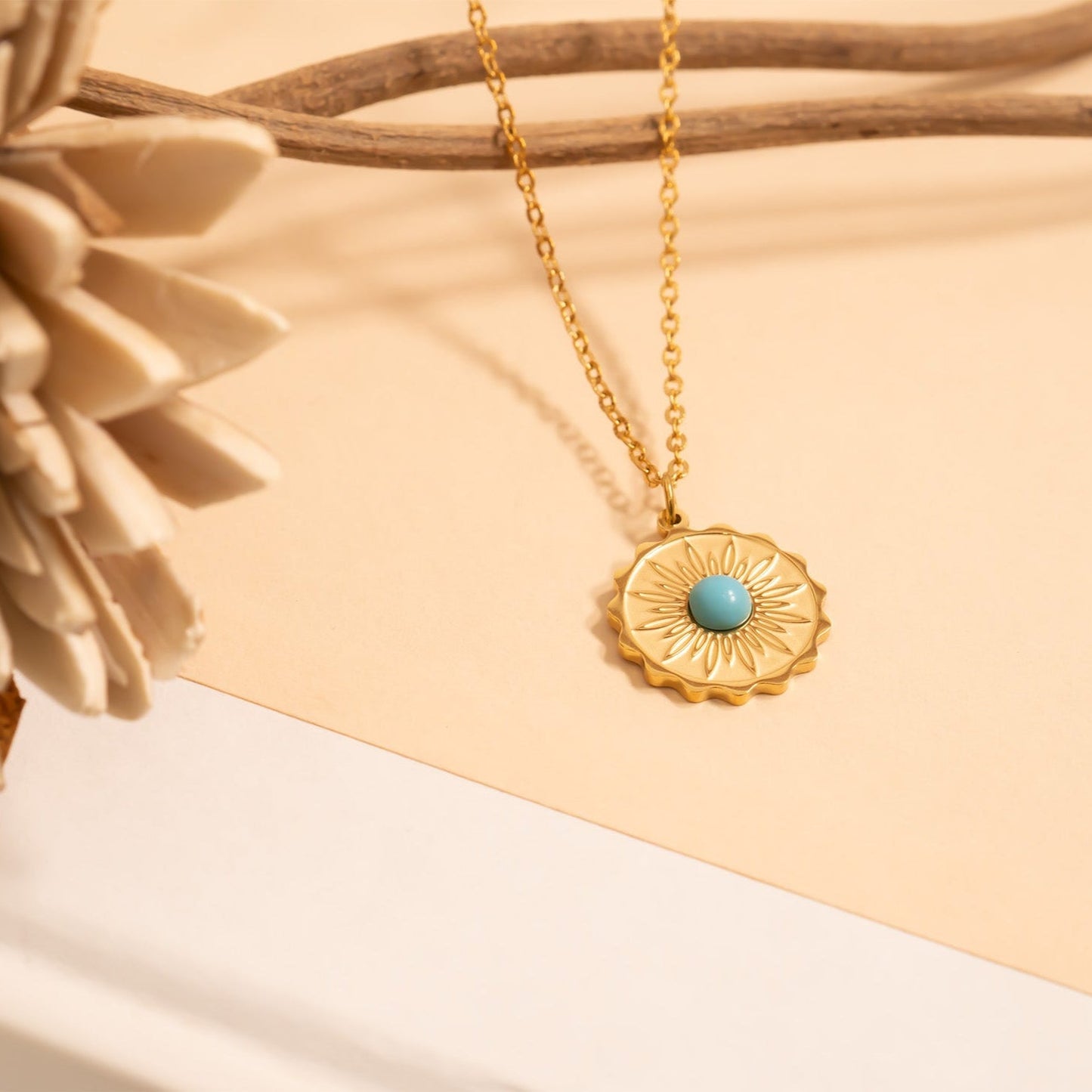 Gold necklace with a gold pendant with flower design and turquoise stone in the middle on a gold chain on a beige background