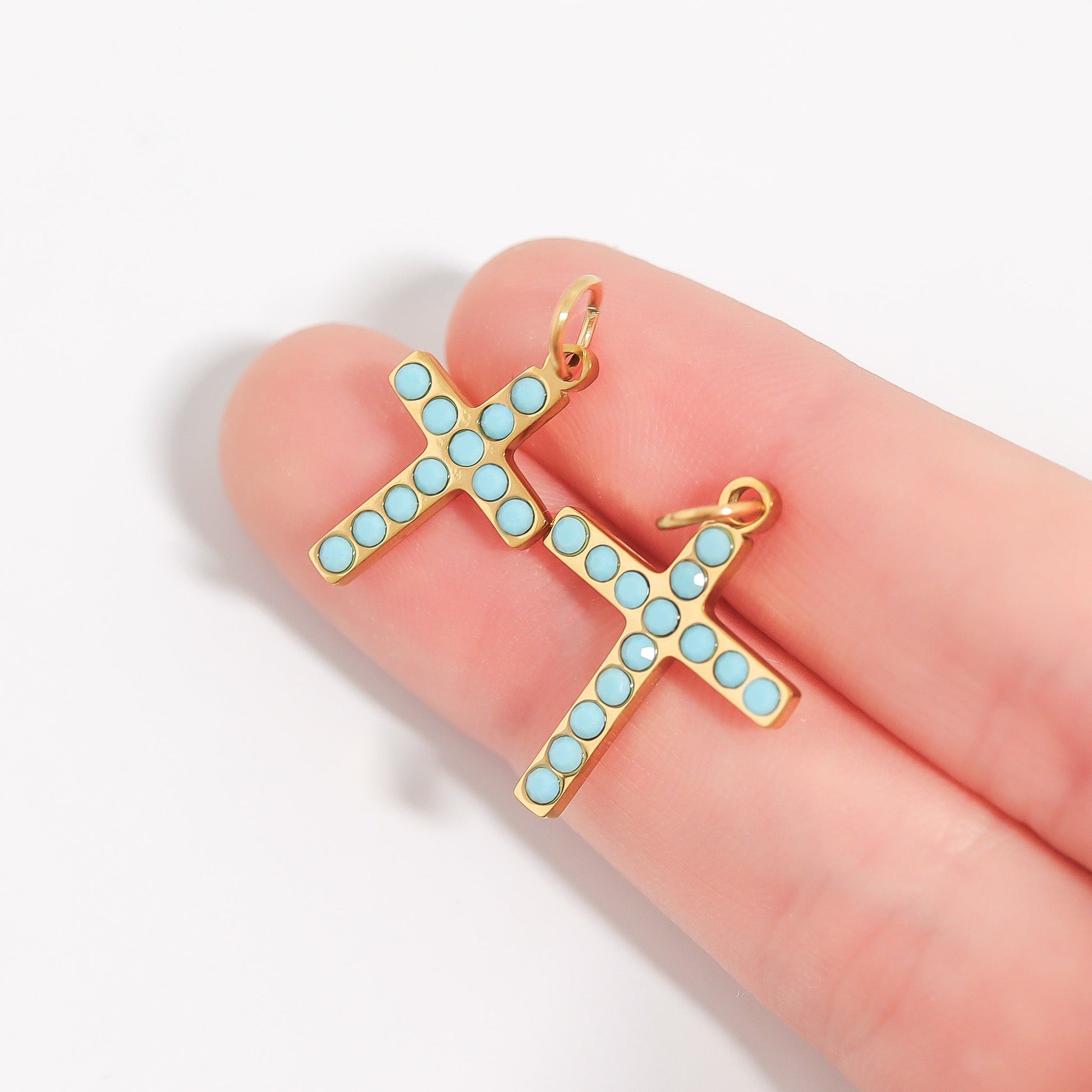 Two gold cross pendants of different sizes with turquoise stones held on two fingers against a white background