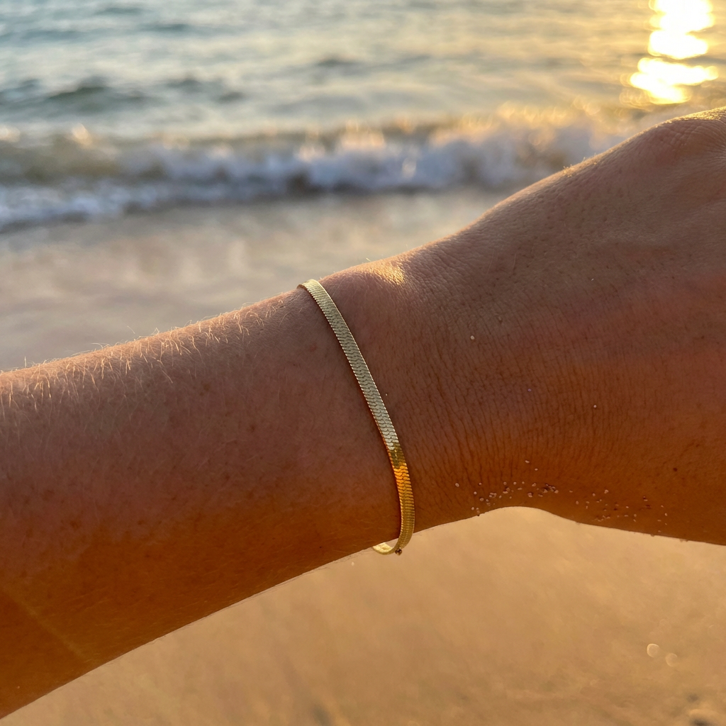 Gold thin herringbone bracelet on a person's wrist with a beach background