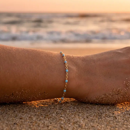 Gold chain bracelet with turquoise beads on a person's wrist at the beach during sunset.