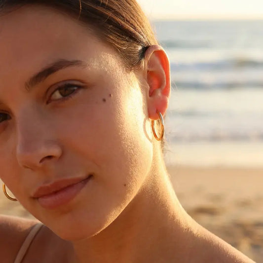 Close-up of a woman's face wearing gold hoop earrings with a blurred beach background