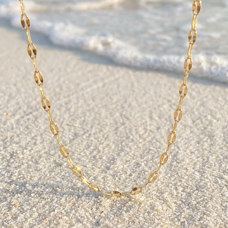 Close up of a gold lip chain necklace on a sandy beach with ocean waves in the background