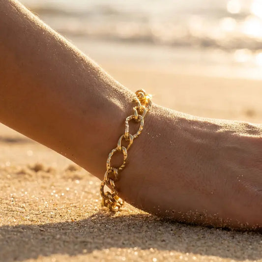 Gold hammered oval chain bracelet on a person's wrist with a beach background