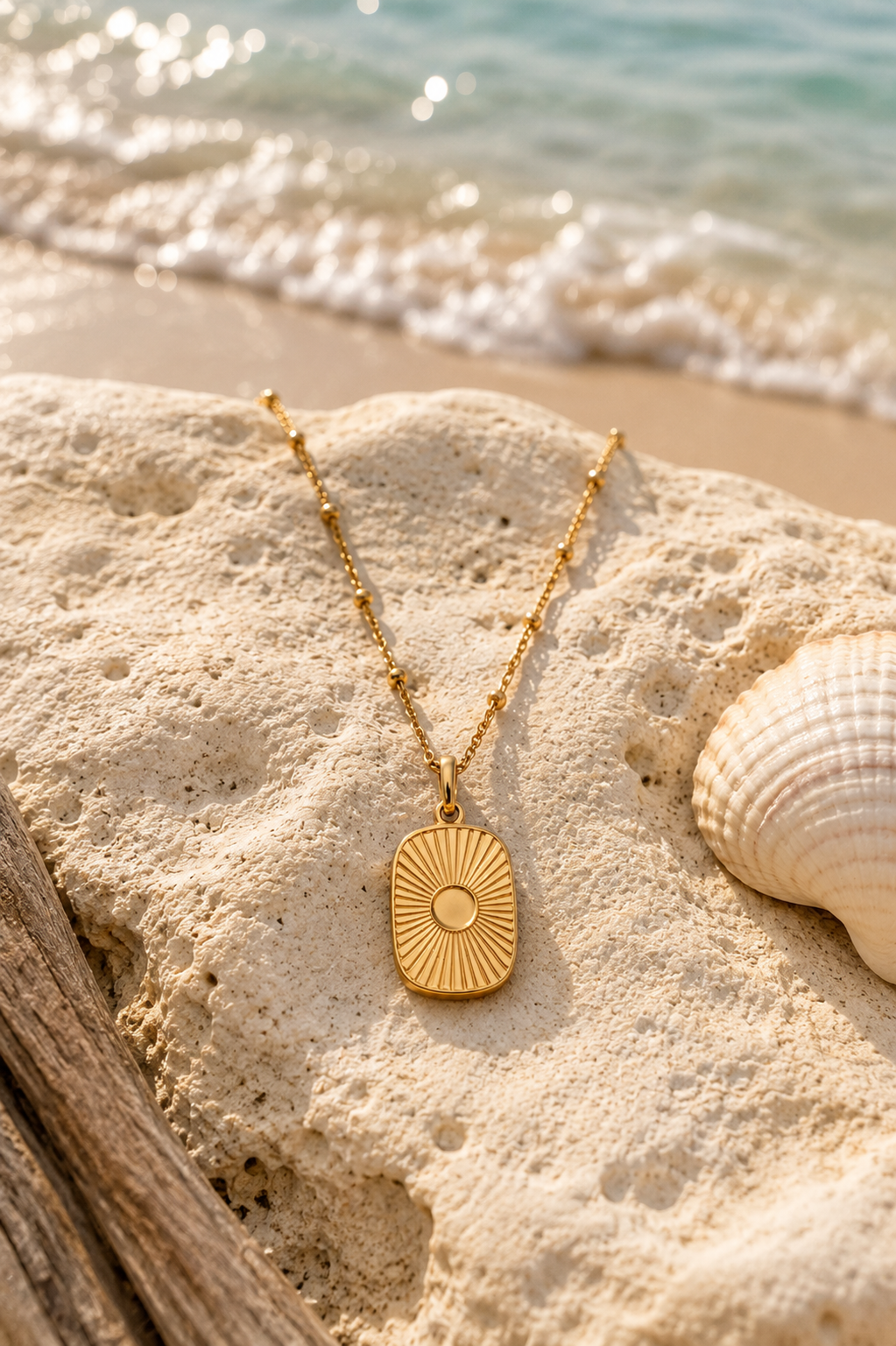Gold satellite chain with sun rays pendant draped over a beach rock next to seashell with crashing ocean wave in the background.