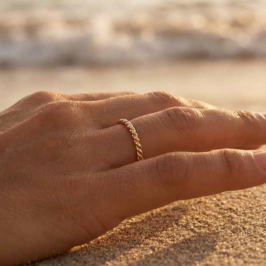 Hand wearing a braided gold ring on a sandy surface with a blurred natural background