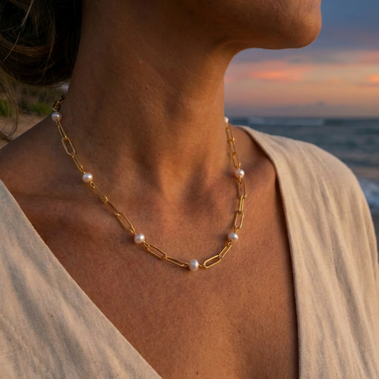Person wearing a gold paperclip chain necklace with pearls between every 2 links against a sunset beach backdrop