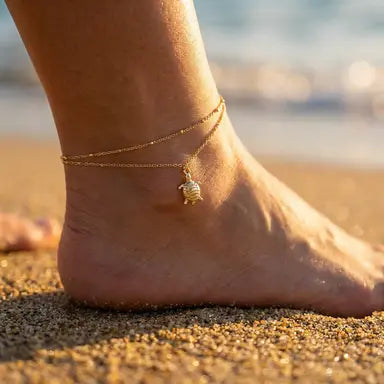Gold layered anklet with sea turtle charm on an ankle in the sand shown from the back side of the foot