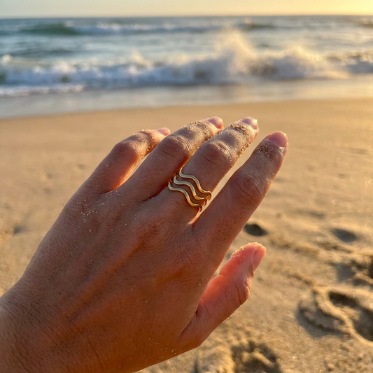 Hand wearing a gold wavy ring on a sandy beach with ocean waves in the background