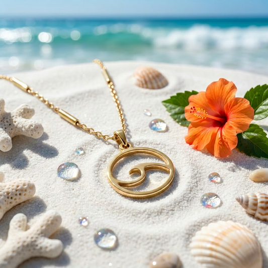 A gold necklace with a circular wave pendant lies on white sand near the ocean, styled with seashells, coral, green leaves, and an orange hibiscus flower.