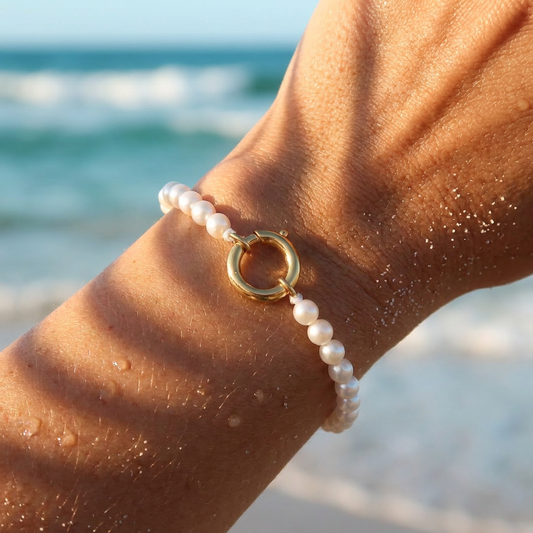 A close-up of a delicate bracelet worn on a person's wrist, featuring small white pearls and a gold ring clasp, set against a blurred beach background with ocean waves.
